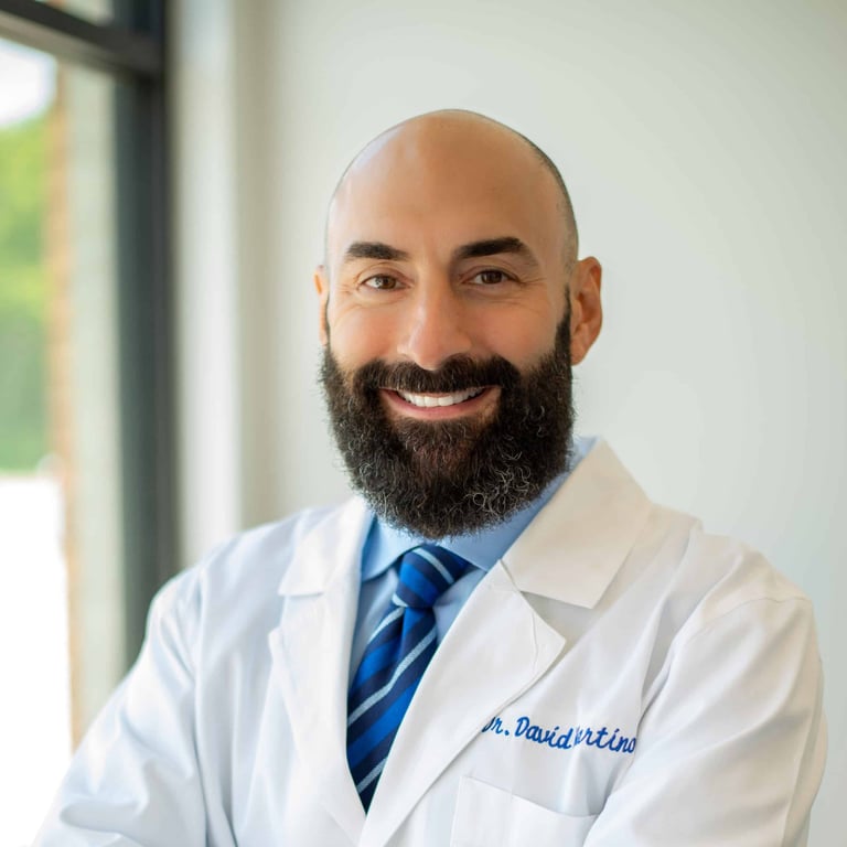 Professional headshot of a male doctor with dark beard wearing white lab coat and blue tie
