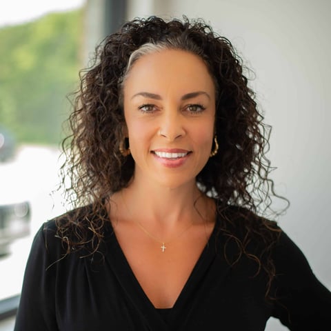 Woman with long curly dark hair wearing a black top with a small cross pendant, smiling at camera in professional portrait