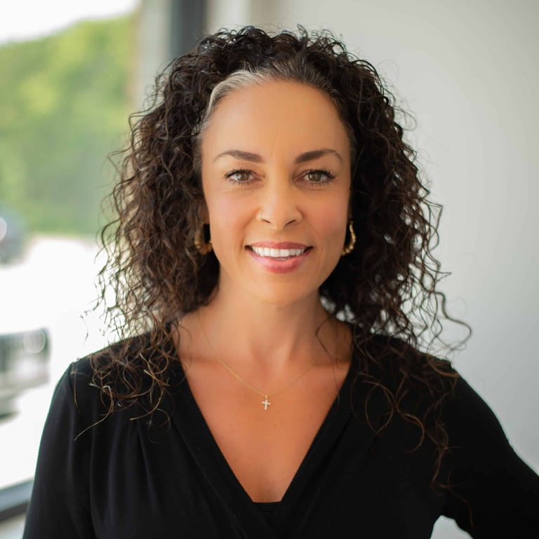 Woman with long curly dark hair wearing a black top with a small cross pendant, smiling at camera in professional portrait